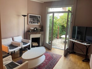 A living room with a fireplace, a television, and a view of the garden through glass doors. - Beautiful house with garden and attic between Marché aux Puces and La Communale (Saint-Ouen-sur-Seine)