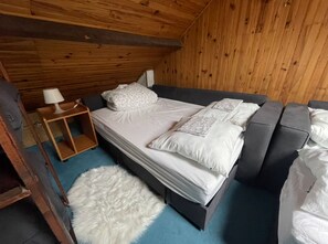 Quarto com teto de madeira, cama com roupa de cama branca, pequena mesa de cabeceira com abajur e tapete branco felpudo.