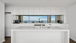 A modern kitchen with a large island, stainless steel appliances, and a view of the sea. - Avoca Views (Avoca Beach)