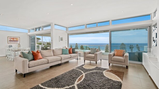 A spacious living room with a large window overlooking the sea, a modern sofa, and a dining area with white chairs. - Avoca Views (Avoca Beach)