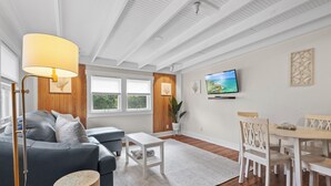 Una sala de estar moderna con un sofá gris, una mesa de centro blanca y una zona de comedor con sillas blancas. Un televisor de pantalla plana está montado en la pared, y hay ventanas con marcos de madera.