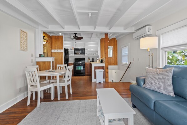 A modern kitchen with white cabinetry, a black refrigerator, and a microwave. There is a dining area with white chairs and a table, and a blue sofa in the living room.