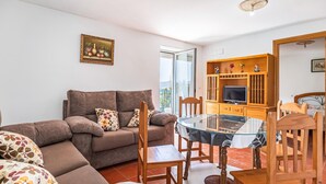 Uma sala de estar com um sofá marrom, almofadas florais, uma mesa de centro de vidro, cadeiras de madeira, uma TV em um armário de madeira e uma obra de arte emoldurada na parede.