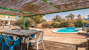 Um pátio externo com piscina, cadeiras azuis, uma mesa e uma pérgula.