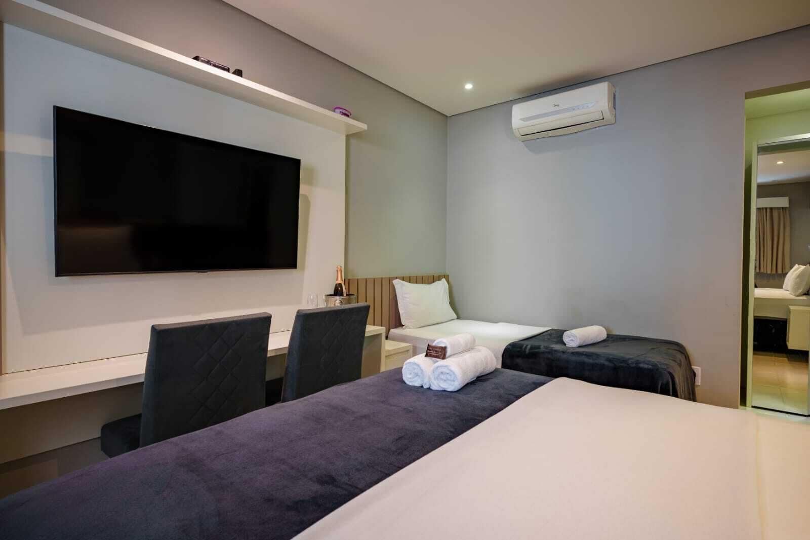 Una habitación de hotel moderna con una televisión de pantalla plana, una cama, un escritorio con sillas y una mesita de noche con una botella de champán.