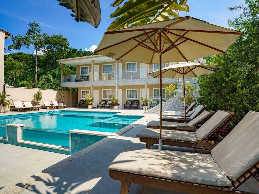 Zona de piscina con sillones de descanso y sombrillas, rodeada de vegetación y un edificio de varios pisos.