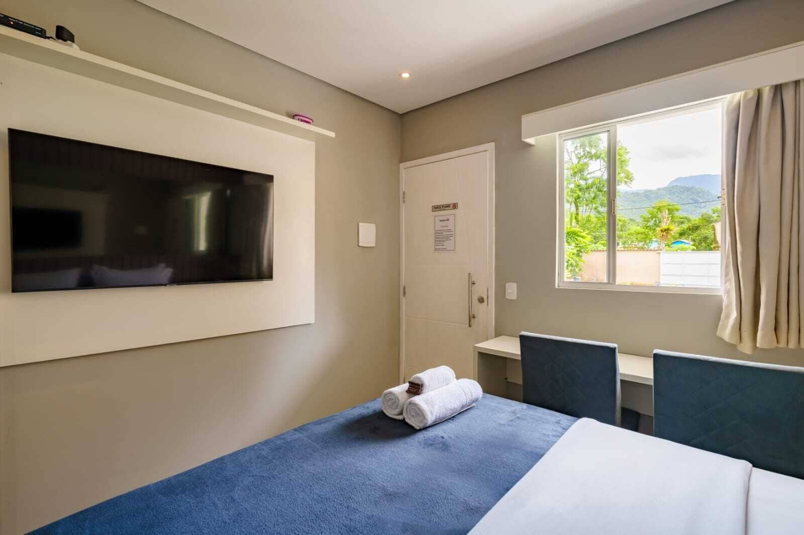 A hotel room with a bed, a desk, a TV, and a window with a view of greenery.