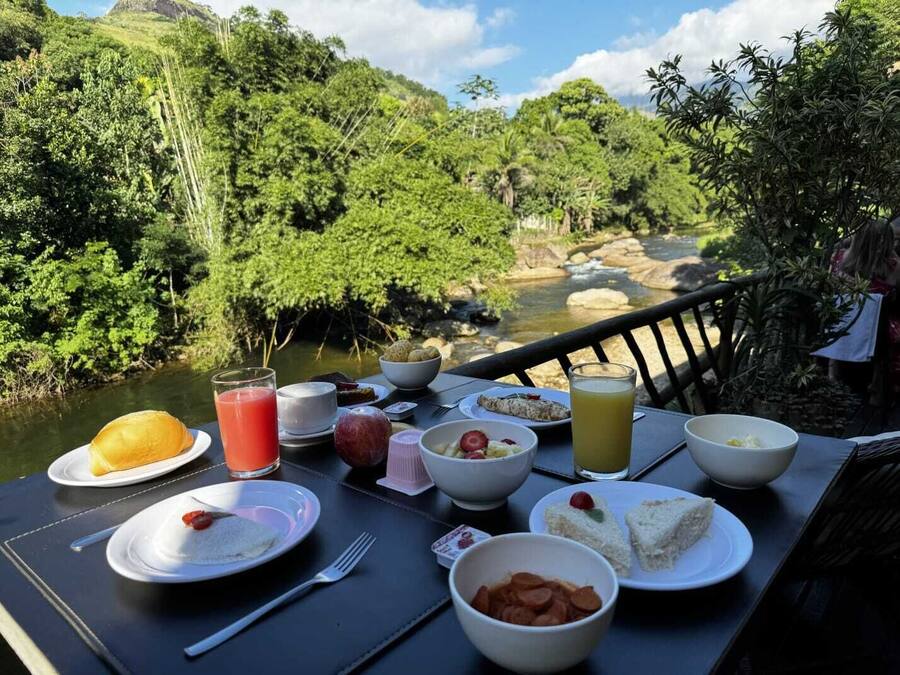 Mesa dispuesta con alimentos para el desayuno, como huevos, tostadas y fruta, con vista a un río y abundante vegetación.