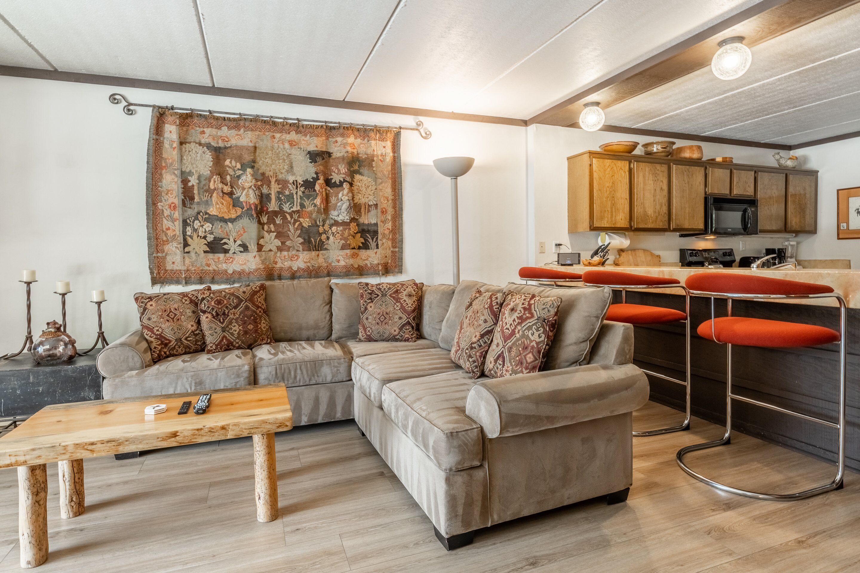 A modern living room with a sectional sofa, a wooden coffee table, and a kitchen area with wooden cabinets and a bar stool.