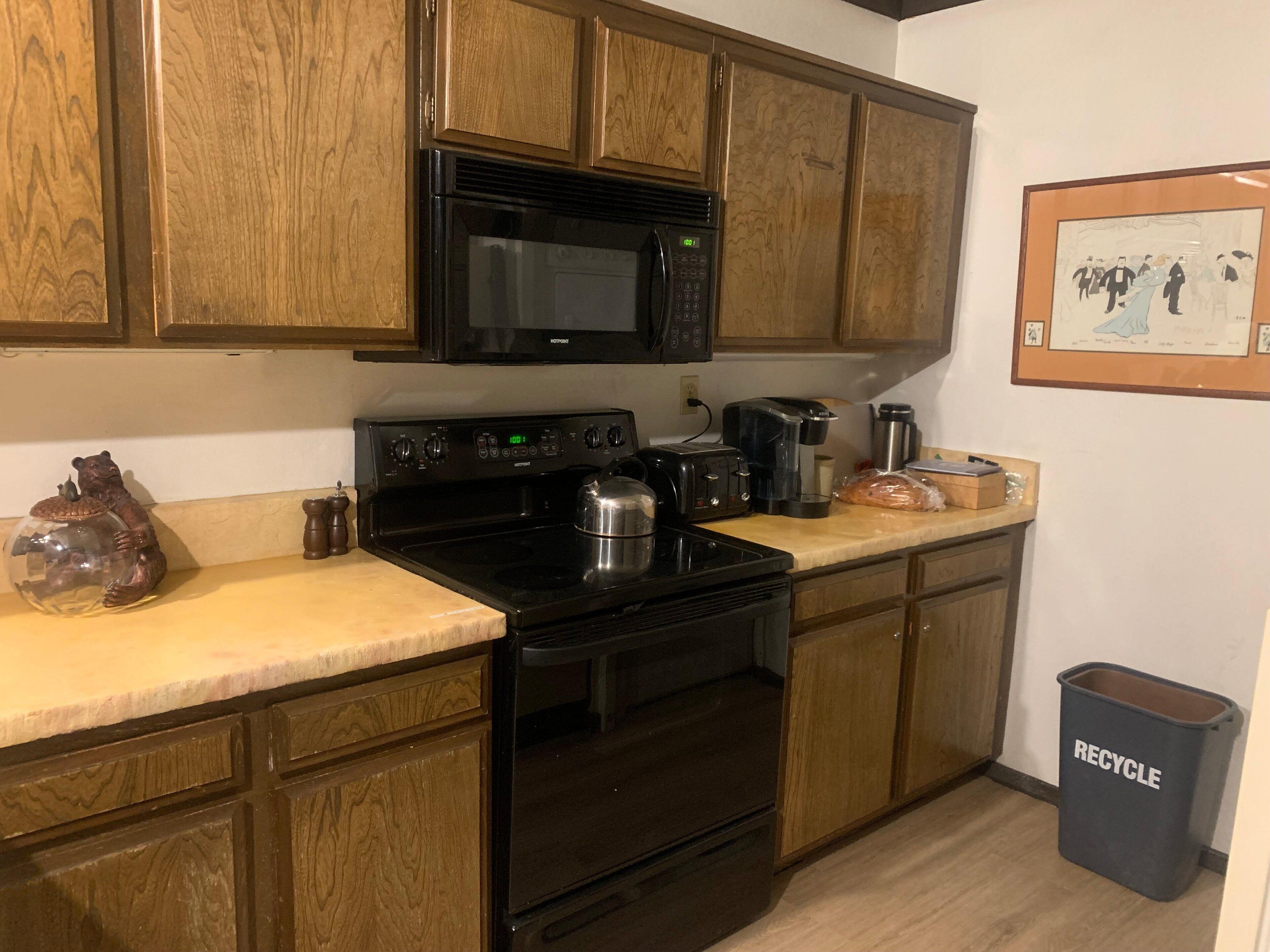 A kitchen with wooden cabinets, a black microwave, a stove, a toaster, and a coffee maker. There's a trash bin labeled "RECYCLE" and a framed picture on the wall.