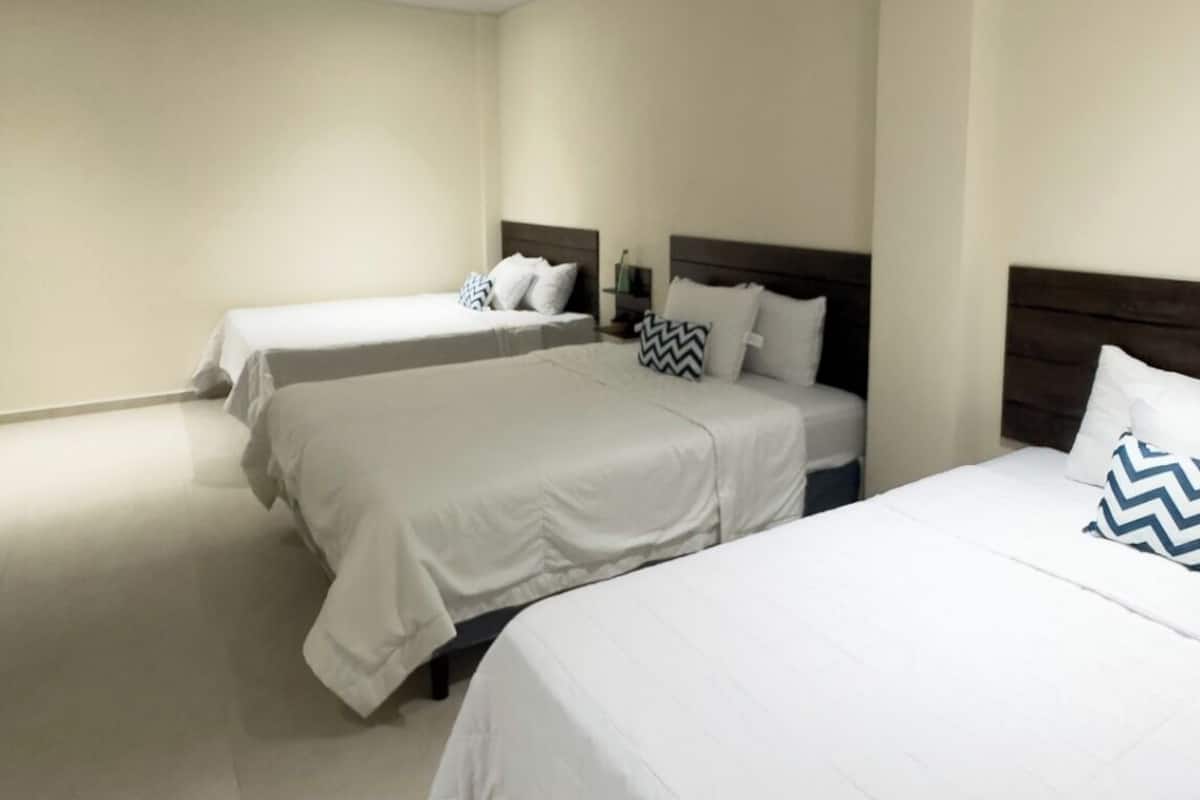 Quarto de hotel com três camas de solteiro, cada uma com cabeceira de madeira e roupa de cama branca.