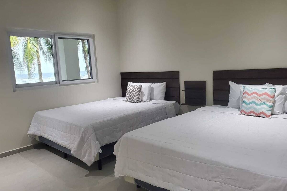Quarto de hotel com duas camas, uma janela grande e vista para palmeiras.