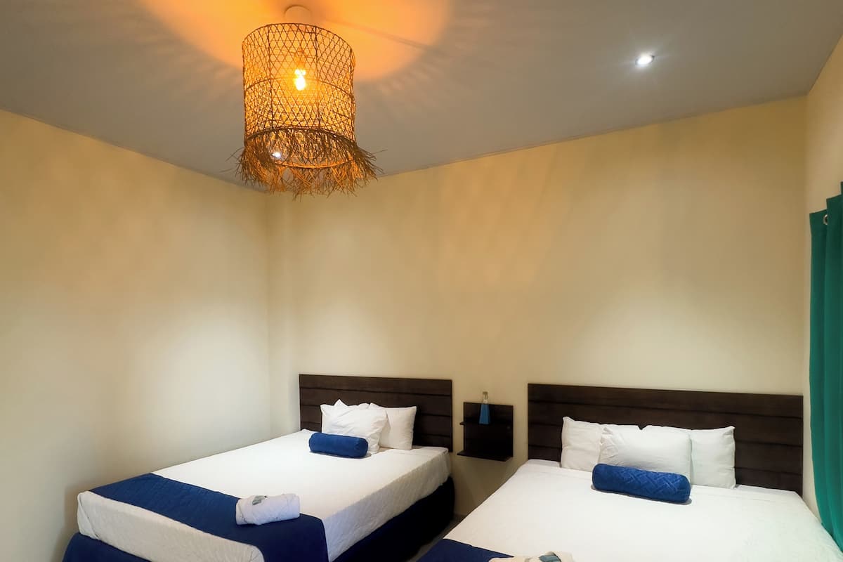 Quarto de hotel com duas camas, cada uma com colcha branca e azul, cabeceira de madeira e luminária pendente.