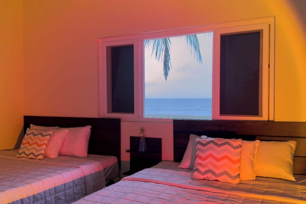 Quarto de hotel com duas camas, uma janela com vista para o oceano e uma mesa de cabeceira com abajur.