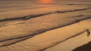 Una playa con el sol poniéndose sobre el océano, dos personas caminando por la orilla.