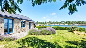 A stone building with a blue door and windows, surrounded by well-maintained grass and shrubs, with a view of a body of water and distant trees. - La petite Pen Er Hoët by Interhome (La Trinité Sur Mer)