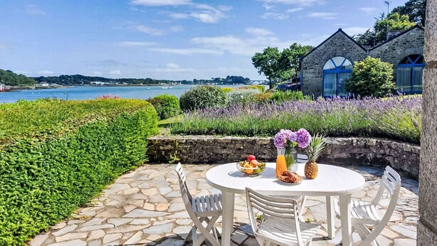 A round table with chairs on a stone patio overlooking a body of water, with a stone building and lush greenery in the background. - La petite Pen Er Hoët by Interhome (La Trinité Sur Mer)
