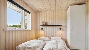 Un dormitorio con paredes de madera, una ventana, una cama con ropa de cama 'LUX', un estante con objetos decorativos y un armario blanco.