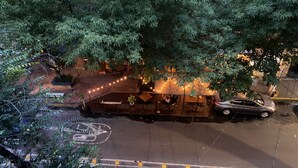 Une vue de rue avec une voiture garée, un bâtiment doté d’un toit en pente et un arbre au premier plan.