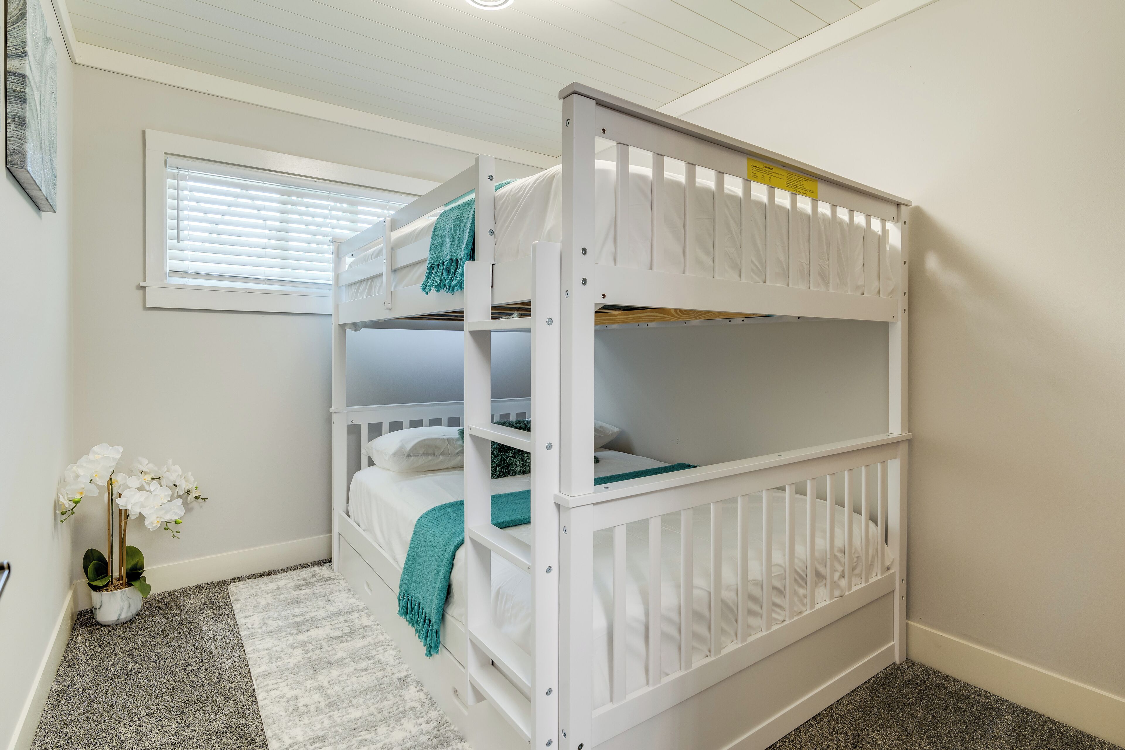 A bunk bed with white bedding and a teal throw blanket. There is a window with blinds, a white door, and a small potted plant with white flowers.