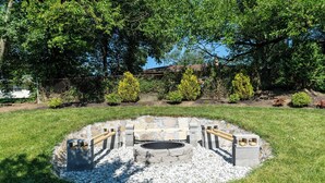 Un foyer circulaire en pierre, agrémenté de bancs, entouré de galets blancs et d’herbe, avec des arbres et une clôture en arrière-plan.