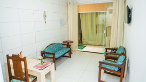 A small, clean room with a tiled floor, two wooden chairs with blue cushions, a small round table with a book and pen, and a glass door leading to a balcony with a green mat. - Pousada Cairu  (Cairu)