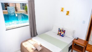 A bedroom with a bed, a small bedside table, a window with a view of a swimming pool, and a colorful pillow. - Pousada Cairu  (Cairu)