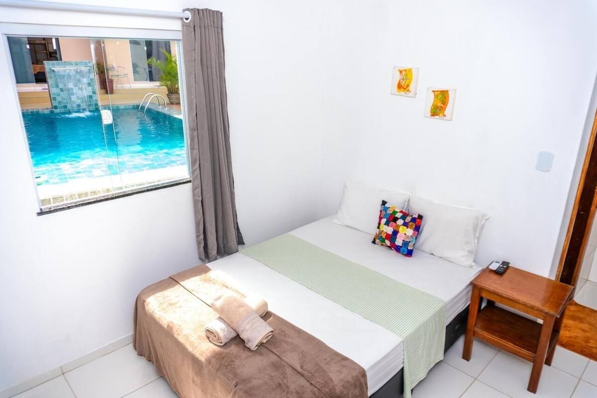 Ein Schlafzimmer mit einem Bett, einem kleinen Nachttisch, einem Fenster mit Blick auf einen Swimmingpool und einem farbenfrohen Kissen.