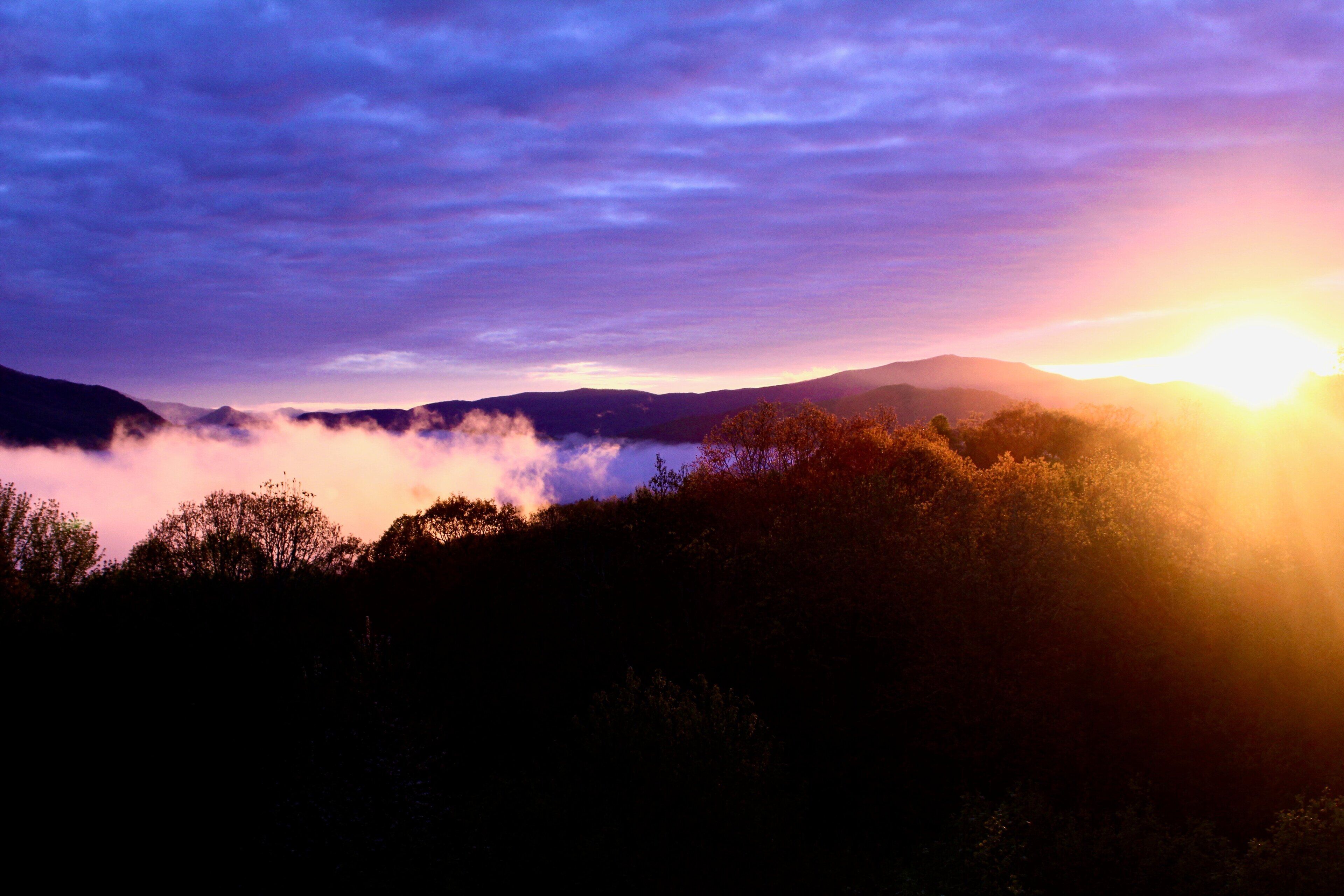 Paisaje montañoso con amanecer o atardecer, con nubes y árboles silueteados.