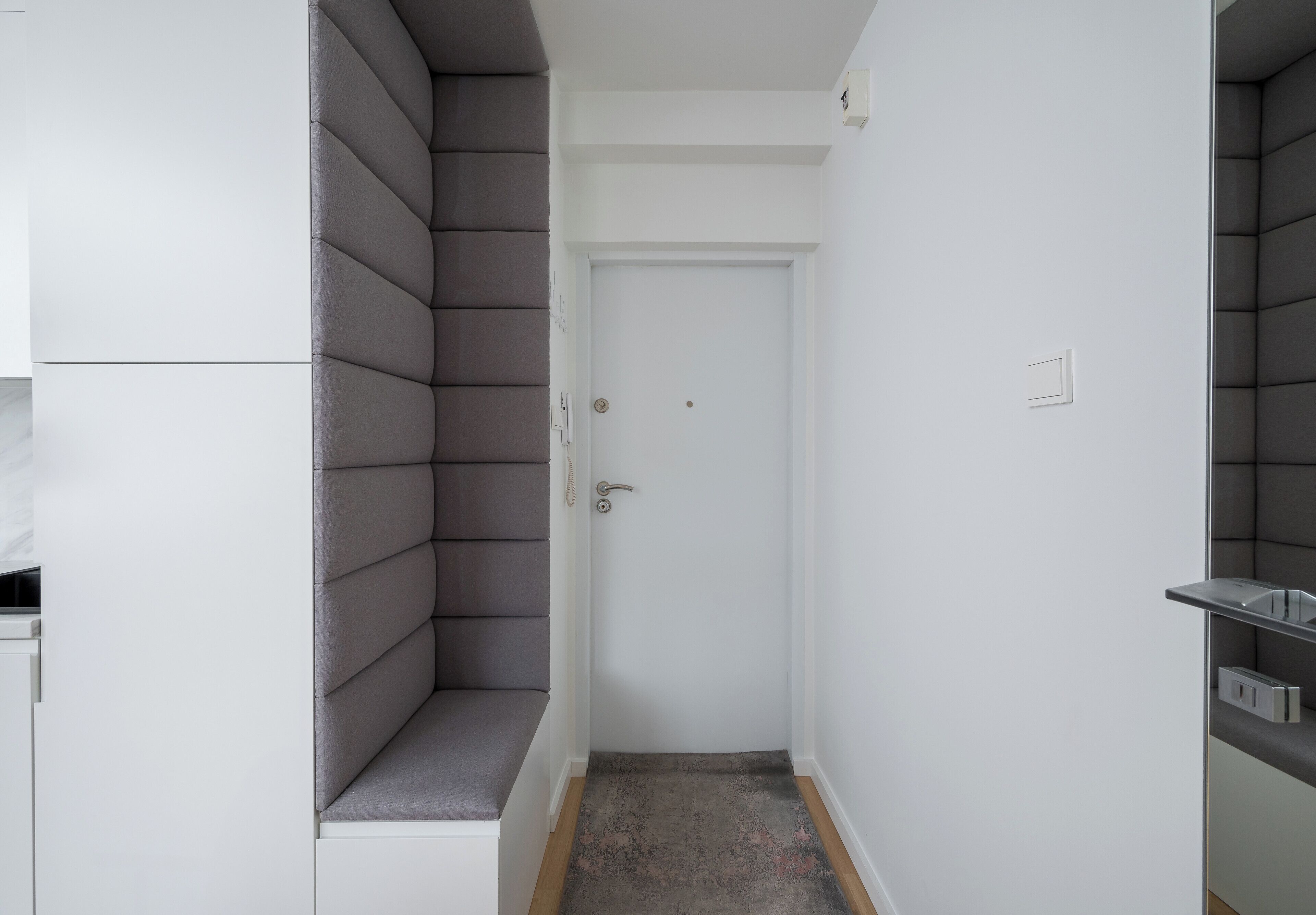 Un pasillo estrecho con una puerta blanca, paredes acolchadas grises y un pequeño banco.