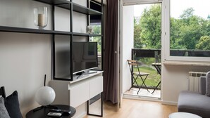 Un salon moderne avec un téléviseur à écran plat, une petite table avec une lampe, un canapé gris et un balcon offrant une vue sur les arbres et un immeuble.