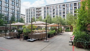 An outdoor patio area with tables and chairs, surrounded by greenery and modern buildings.