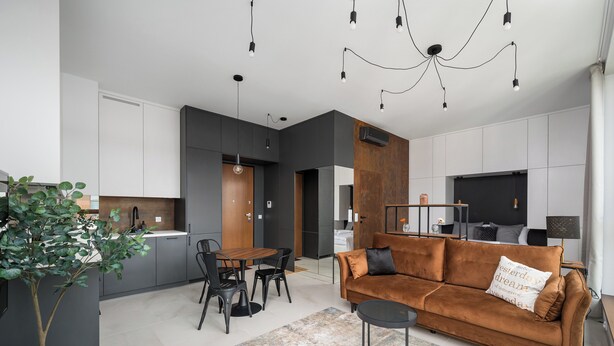 A modern living room with a brown sofa, a dining table, and chairs. There is a kitchen area with cabinets and a refrigerator.