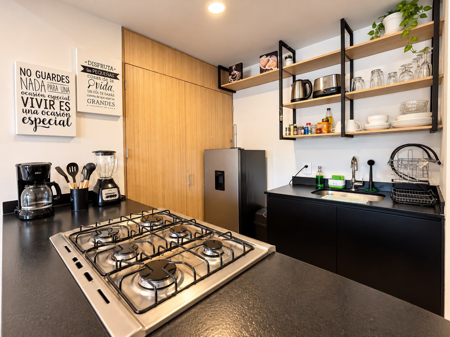 A modern kitchen with a gas stove, coffee maker, blender, and a refrigerator. There are wooden cabinets and a black countertop.