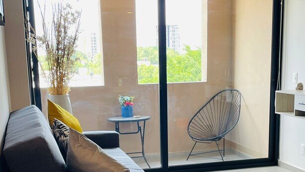 A modern living room with a sofa, a small round table, and a chair. There is a sliding glass door leading to a balcony with a view of buildings and trees. - Arcos Vallarta 2be/2ba/1parking (Guadalajara)