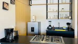 A modern kitchen with a gas stove, a coffee maker, a refrigerator, and a shelf with glassware and kitchen utensils. - Arcos Vallarta 2be/2ba/1parking (Guadalajara)