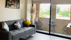 A living room with a grey sofa, a small round table, and a chair. There is a large window with a view of buildings and trees. - Arcos Vallarta 2be/2ba/1parking (Guadalajara)