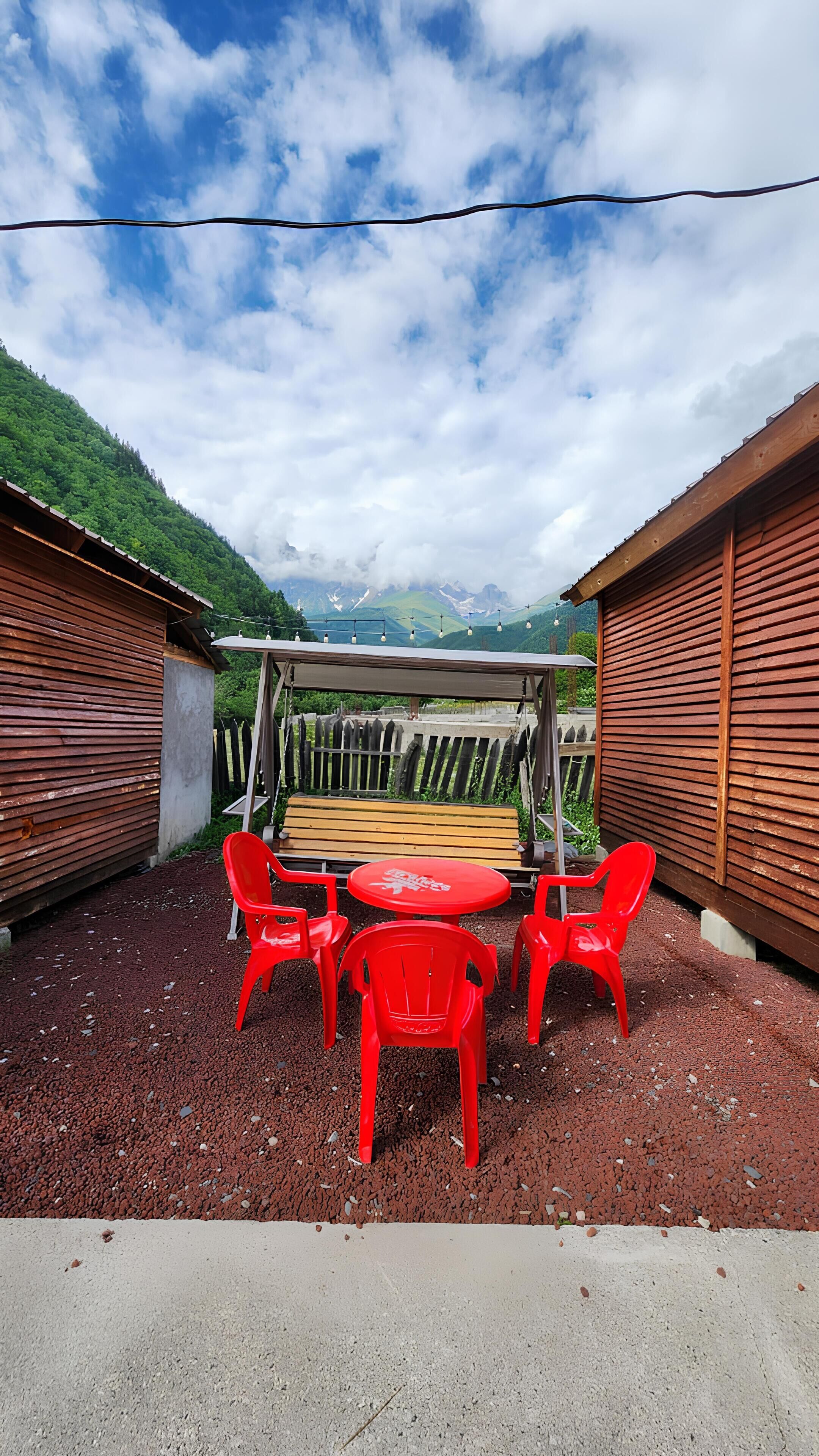 Un espace extérieur aménagé avec des chaises en plastique rouge et une table, entour é de bâtiments en bois et offrant une vue sur les montagnes.