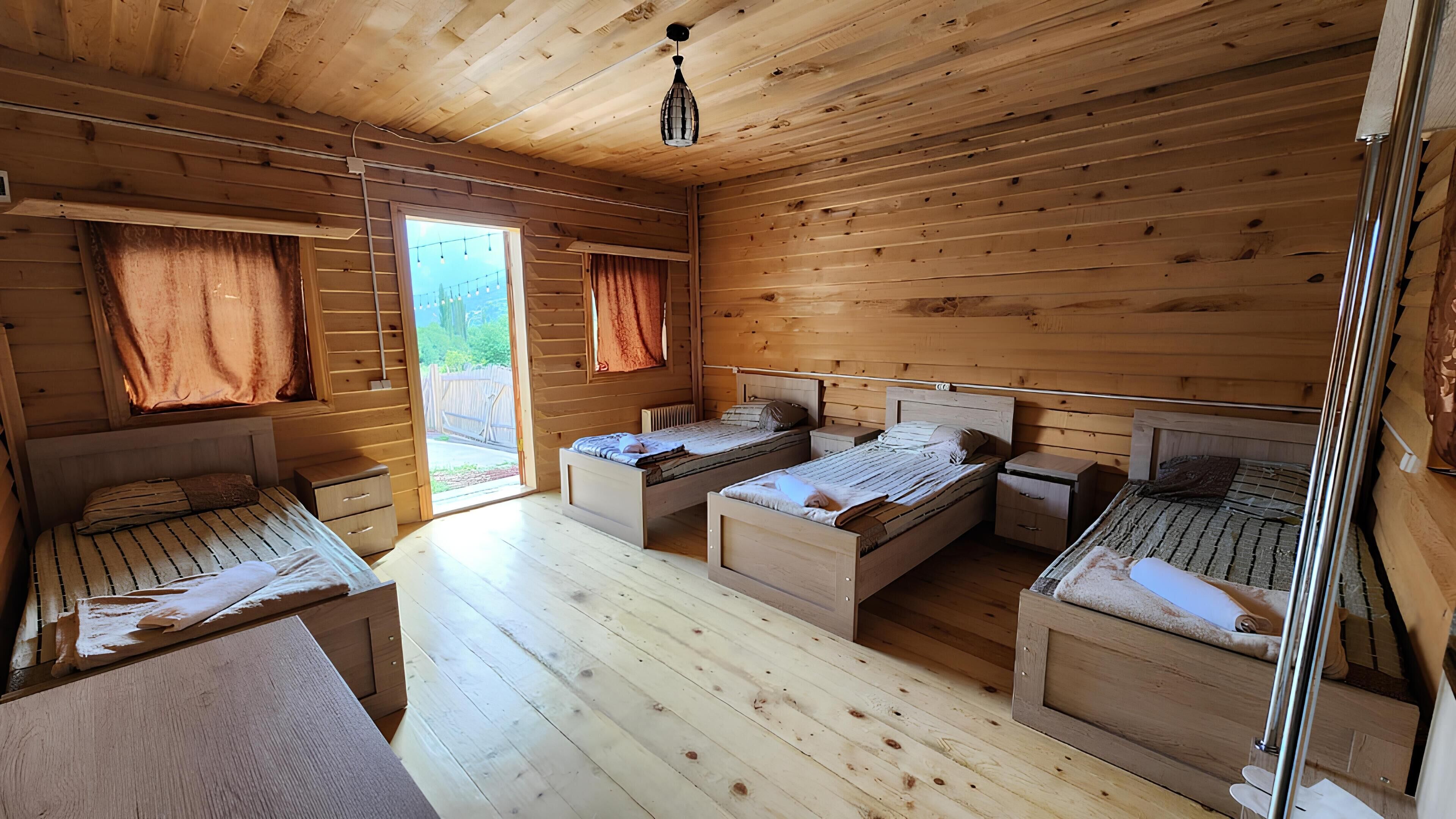 A cabin with four wooden beds arranged in a U-shape, a wooden floor, and a large window with curtains.