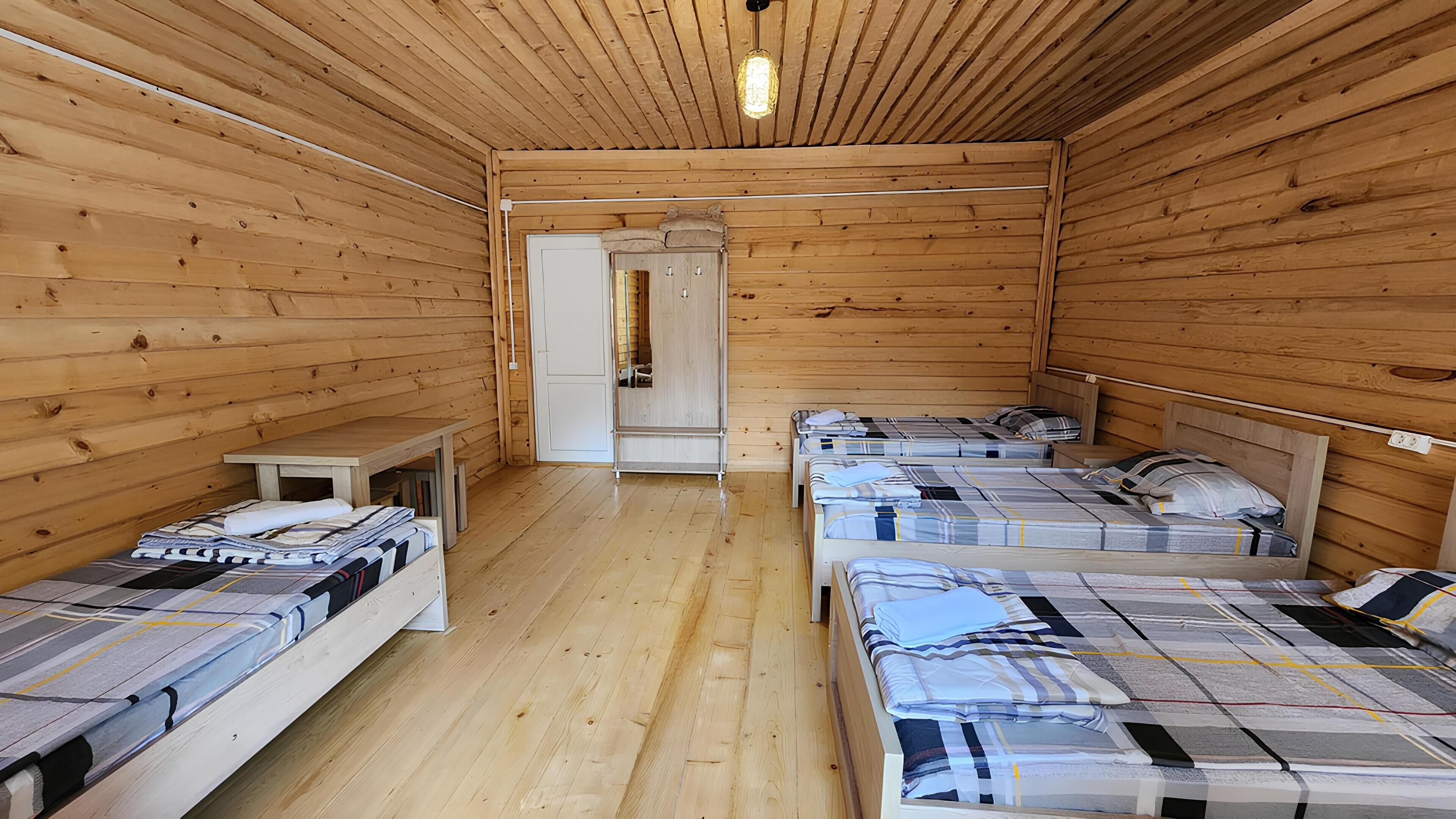 Uma cabana com paredes e pisos de madeira, duas camas de solteiro, uma mesa pequena e um guarda-roupa.