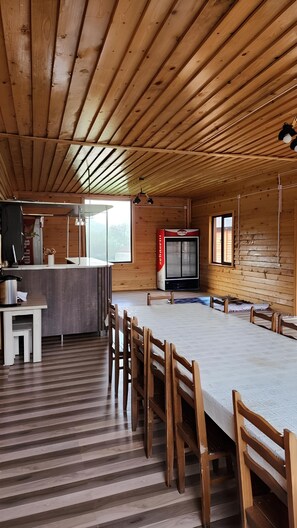 A spacious wooden cabin with a long dining table, wooden chairs, a kitchen area, and a large window providing natural light. - Ushba Gate House (Mestia)