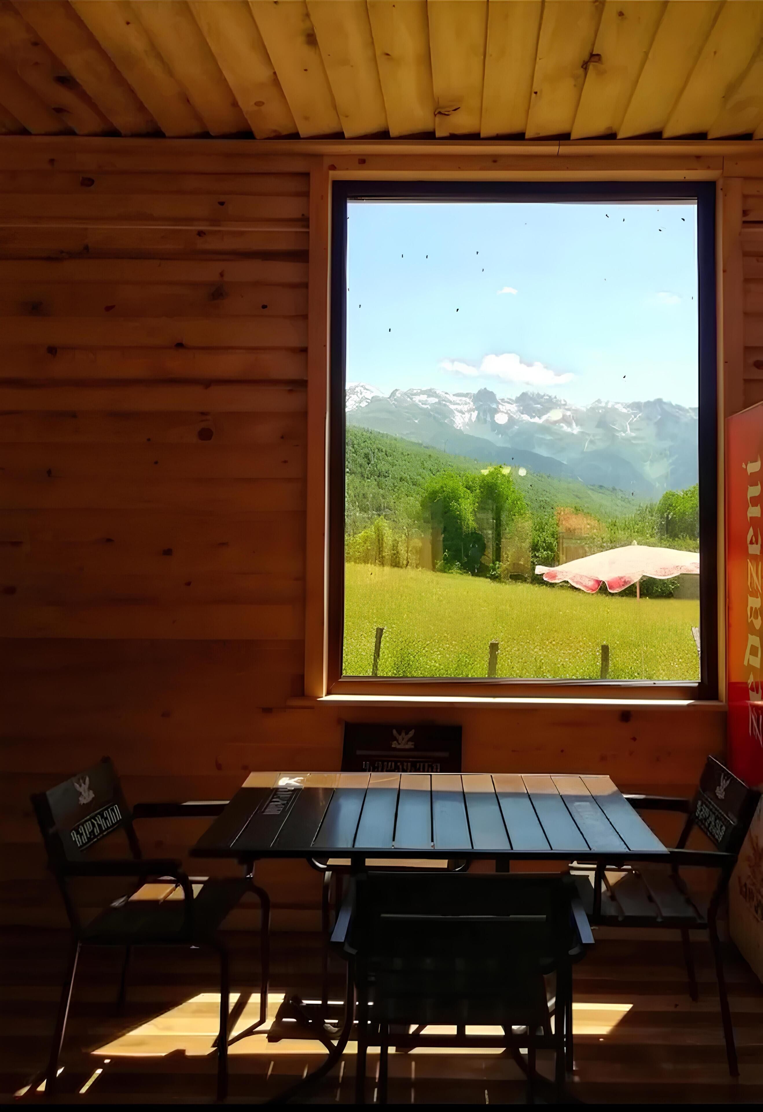 Una cabaña de madera con un ventanal grande que da a un campo verde y montañas. En el interior hay una mesa y dos sillas.