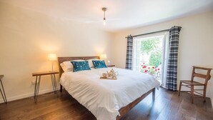 A bedroom with a wooden bed, two bedside tables, a window with curtains, and a view of greenery outside. - Olive Cottage, Wickham Market (Wickham Market)