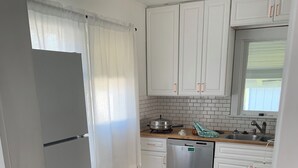 Una cocina con armarios blancos, lavavajillas de acero inoxidable y una ventana con cortinas translúcidas.