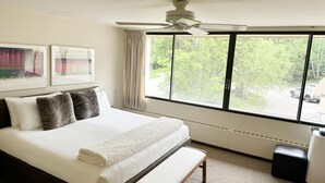 Ein Schlafzimmer mit einem großen Bett, einem Deckenventilator, einem Fenster mit Blick auf Bäume und einem abgestellten Lastwagen, sowie einer kleinen Bank.