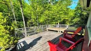 A wooden deck with a railing, surrounded by lush green trees. - Serene & Cozy 3BR Home w/ Large Deck on River (Stowe)