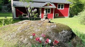 A red house with a stone stack in front, surrounded by a grassy yard and tulips. - Serene & Cozy 3BR Home w/ Large Deck on River (Stowe)