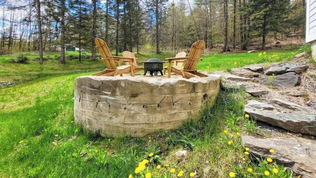 A stone fire pit with two Adirondack chairs and a table, surrounded by a grassy area with yellow flowers and a forest in the background. - Clean, perfect home a mile from historic Waterbury (Waterbury Village Historic District)