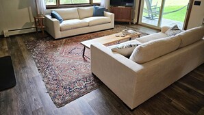 A living room with a beige sofa, a coffee table, a TV, and a large window with a view of greenery. - Rustic Farmstay, Bucolic retreat w/Endless Views (Stowe)