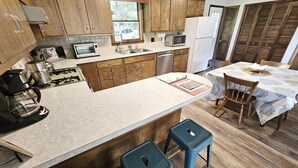 A modern kitchen with wooden cabinets, a central island with stools, a dining table with chairs, and a view of the outside through a window. - Rustic Farmstay, Bucolic retreat w/Endless Views (Stowe)